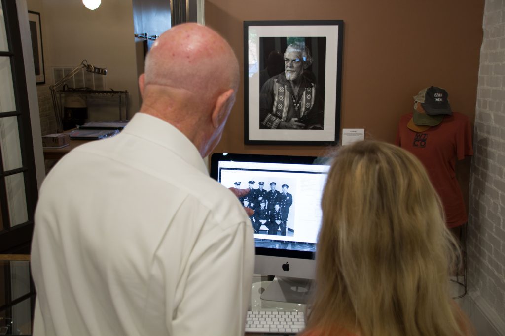 A man pointing to a black and white photo on the screen of an iMac to identify police officers pictured in the image as a female employee of the W7thCo Gallery takes notes.